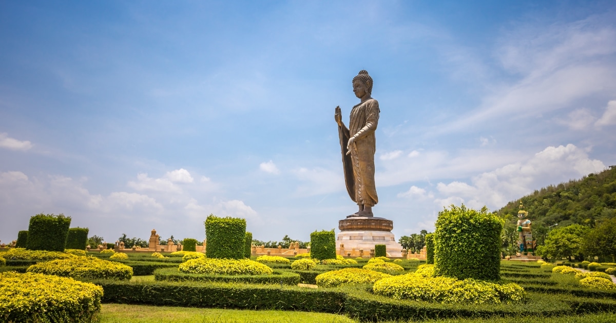 พระพุทธรูปขนาดใหญ่ตั้งตระหง่านอยู่ท่ามกลางสวนที่ตกแต่งอย่างประณีตและพุ่มไม้ที่ตัดแต่งอย่างสวยงาม ทำให้ที่นี่เป็นสถานที่ท่องเที่ยวในกาญจนบุรีที่เงียบสงบภายใต้ท้องฟ้าสีครามและเมฆที่ลอยอยู่ประปราย.