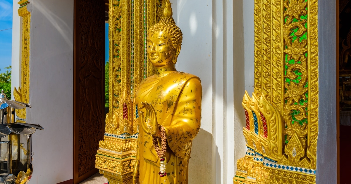 พระพุทธรูปทองคำพร้อมพวงมาลัยตั้งอยู่หน้าทางเข้าวัดที่ประดับประดาอย่างวิจิตรด้วยลวดลายทองคำอันประณีต เป็นไฮไลท์ที่ต้องชมเมื่อมาเที่ยวกาญจนบุรี.