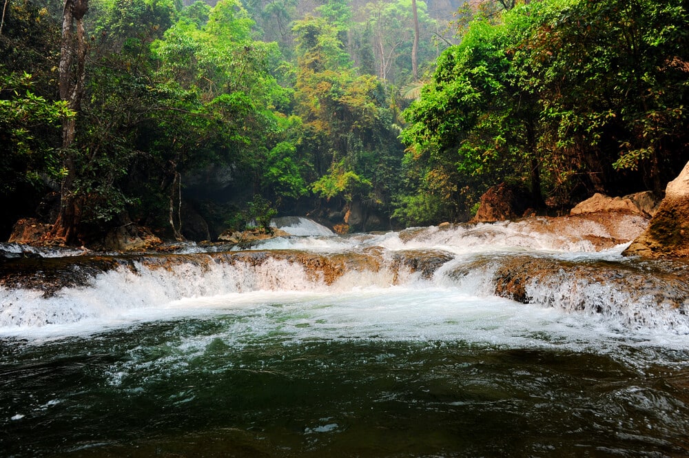 แม่น้ำไหลผ่านโขดหินในป่าฝนอันเขียวชอุ่ม ล้อมรอบด้วยต้นไม้เขียวขจีและพืชพรรณหนาแน่น—เหมาะสำหรับผู้ที่ต้องการสำรวจสถานที่ท่องเที่ยวในกาญจนบุรีและดื่มด่ำกับความงดงามของสถานที่ท่องเที่ยวในกาญจนบุรี.