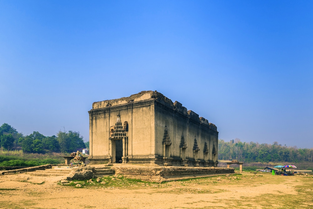 อาคารหินรูปสี่เหลี่ยมผืนผ้าที่ผ่านกาลเวลา มีหน้าต่างโค้งตั้งอยู่โดดเดี่ยวบนพื้นทรายใต้ท้องฟ้าสีฟ้าใส—จุดที่น่าสนใจสำหรับผู้ที่กำลังสำรวจสถานที่ท่องเที่ยวในกาญจนบุรี.