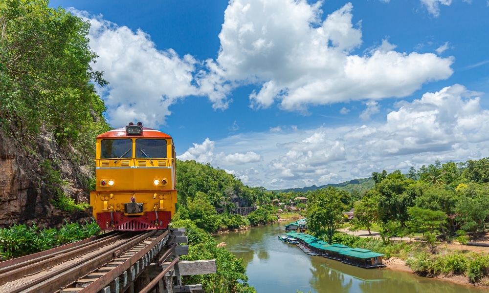 รถไฟสีเหลืองข้ามสะพานเหนือแม่น้ำที่ล้อมรอบด้วยต้นไม้สีเขียวใน กาญจนบุรีที่เที่ยว โดยมีเรือจอดอยู่ตามริมฝั่งแม่น้ำและท้องฟ้าที่มีเมฆครึ้มอยู่เหนือศีรษะ.