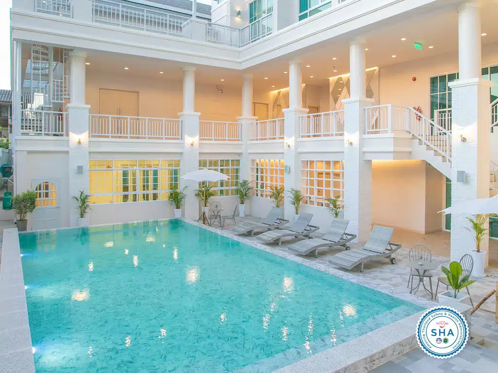 โรงแรมเชียงรายสมัยใหม่ที่มีลานกลางแจ้งพร้อมสระว่ายน้ำกลางแจ้งรูปสี่เหลี่ยมผืนผ้า เก้าอี้เลานจ์หลายตัว ร่ม และสถาปัตยกรรมสองชั้นสีขาว โลโก้การรับรอง SHA มองเห็นได้ชัดเจนที่มุม.