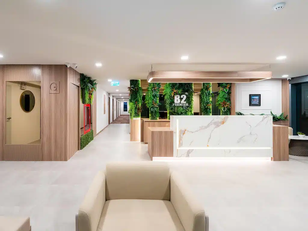 ล็อบบี้โรงแรมสมัยใหม่พร้อมที่นั่งสีเบจ เคาน์เตอร์ต้อนรับหินอ่อน ผนังตกแต่งด้วยต้นไม้สีเขียว และของตกแต่งแบบมินิมอล เหมาะสำหรับนักเดินทางที่กำลังมองหา โรงแรมเชียงใหม่ ที่มีสไตล์ ทางเดินทอดยาวไปทางซ้ายของบริเวณต้อนรับ.