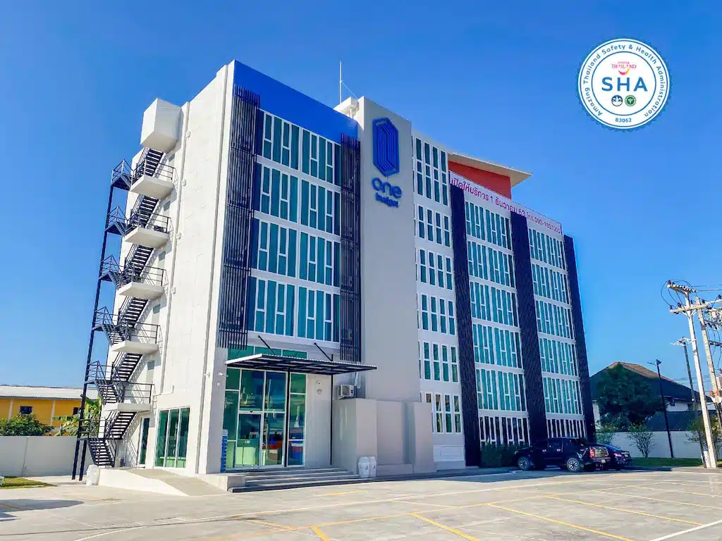 โรงแรมสี่ชั้นสมัยใหม่ในเชียงราย พร้อมแผงสีน้ำเงินและขาว บันไดภายนอก และลานจอดรถขนาดใหญ่ภายใต้ท้องฟ้าสีครามสดใส โลโก้การรับรอง SHA ปรากฏอยู่ที่มุมขวาบน.