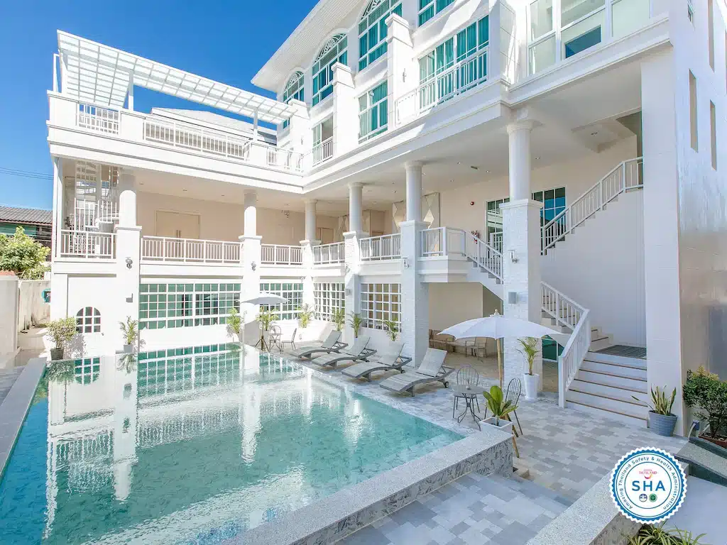 อาคารสีขาวสมัยใหม่พร้อมหน้าต่างบานใหญ่และระเบียง มีสระว่ายน้ำกลางแจ้งล้อมรอบด้วยเก้าอี้อาบแดดและร่มใต้ท้องฟ้าสีฟ้าใส—เหมาะสำหรับการเข้าพักที่ โรงแรมเชียงราย ของคุณ.