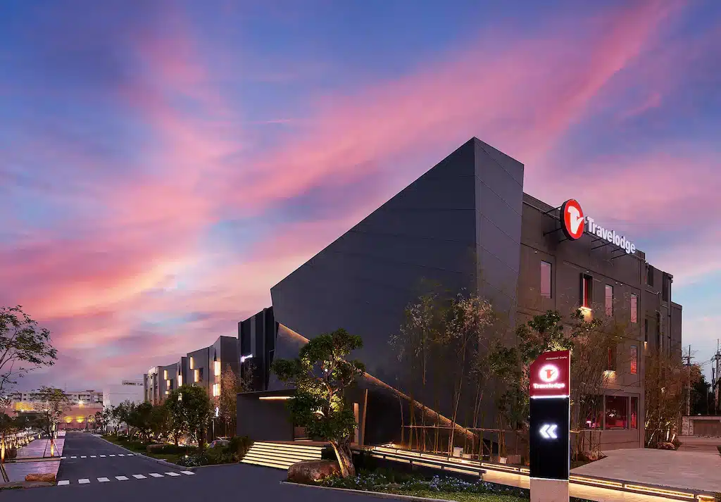 อาคารโรงแรม Travelodge ทันสมัยที่มีสถาปัตยกรรมเหลี่ยมมุมในยามพระอาทิตย์ตกดิน พร้อมป้ายไฟส่องสว่างและภูมิทัศน์โดยรอบภายใต้ท้องฟ้าสีชมพูและสีน้ำเงิน—เหมาะสำหรับผู้ที่มองหา โรงแรมเชียงใหม่ ในบรรยากาศเมืองที่มีสไตล์.