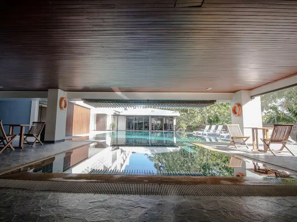 บริเวณสระว่ายน้ำในร่มพร้อมเก้าอี้ไม้ โต๊ะ และห่วงยางสำหรับความปลอดภัย ณ โรงแรมเชียงใหม่ เปิดสู่พื้นที่กลางแจ้งที่มีเก้าอี้เลานจ์และต้นไม้ให้เห็นอยู่เบื้องหลัง.