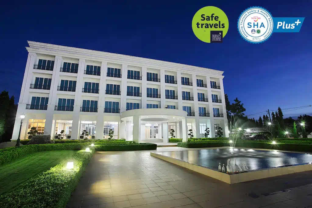 โรงแรมเชียงรายสีขาวสูงสี่ชั้น มีทางเข้าตรงกลาง ถนนใหญ่สำหรับรถยนต์ ต้นไม้ประดับสวยงาม และน้ำพุรูปสี่เหลี่ยมผืนผ้าอยู่หน้าโรงแรม ถ่ายรูปตอนพลบค่ำ แสดงป้าย "เดินทางปลอดภัย" และ "SHA Plus+.