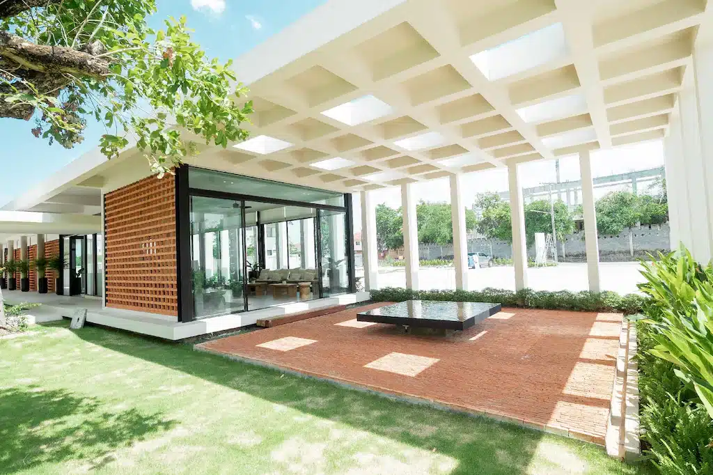 อาคารสมัยใหม่พร้อมประตูกระจกบานใหญ่ หลังคาโครงเหล็กเปิดโล่ง ผนังอิฐ และสระน้ำสะท้อนกลาง ล้อมรอบด้วยพื้นที่สีเขียวและแสงธรรมชาติ—เหมาะอย่างยิ่งสำหรับผู้ที่มองหา โรงแรมในเชียงใหม่ ที่มีสไตล์หรือ ที่พักเชียงใหม่ ที่เงียบสงบ.