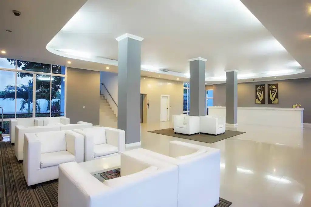 ล็อบบี้โรงแรมเชียงรายสมัยใหม่ พร้อมเก้าอี้แขนสีขาว โซฟา และเสา ตกแต่งด้วยเคาน์เตอร์ต้อนรับ ศิลปะบนผนัง และหน้าต่างบานใหญ่ที่เปิดรับแสงธรรมชาติ.