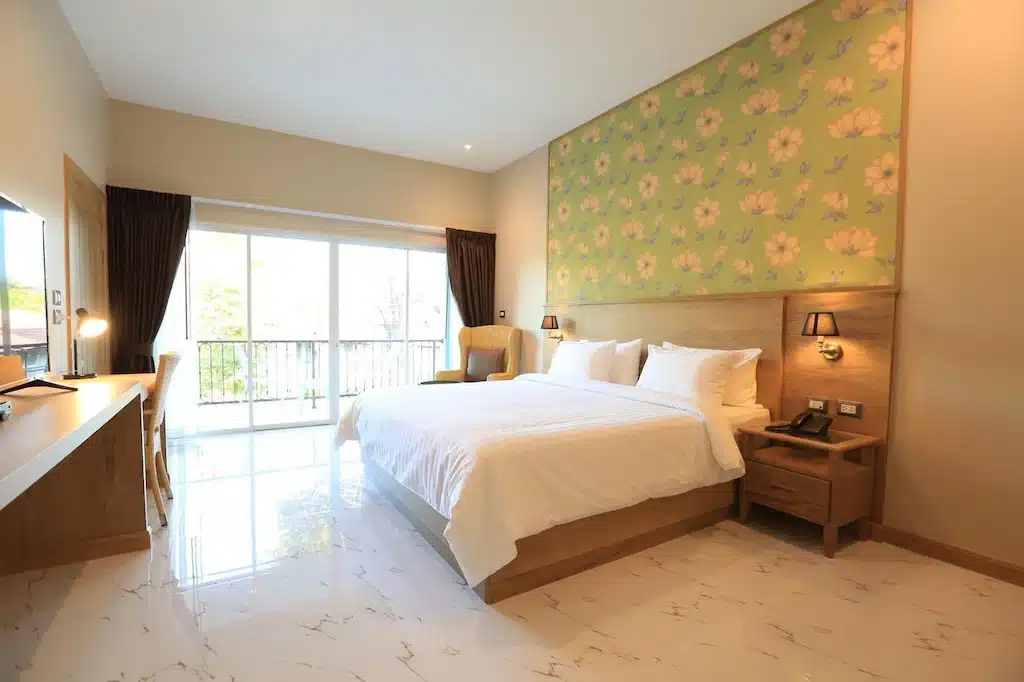 ห้องพักโรงแรมเชียงรายที่สว่างสดใส พร้อมเตียงขนาดใหญ่ ผนังตกแต่งด้วยลวดลายดอกไม้ โต๊ะทำงาน เก้าอี้มีพนักพิง โคมไฟข้างเตียง และหน้าต่างบานใหญ่จากพื้นจรดเพดานพร้อมผ้าม่าน มองเห็นระเบียง.