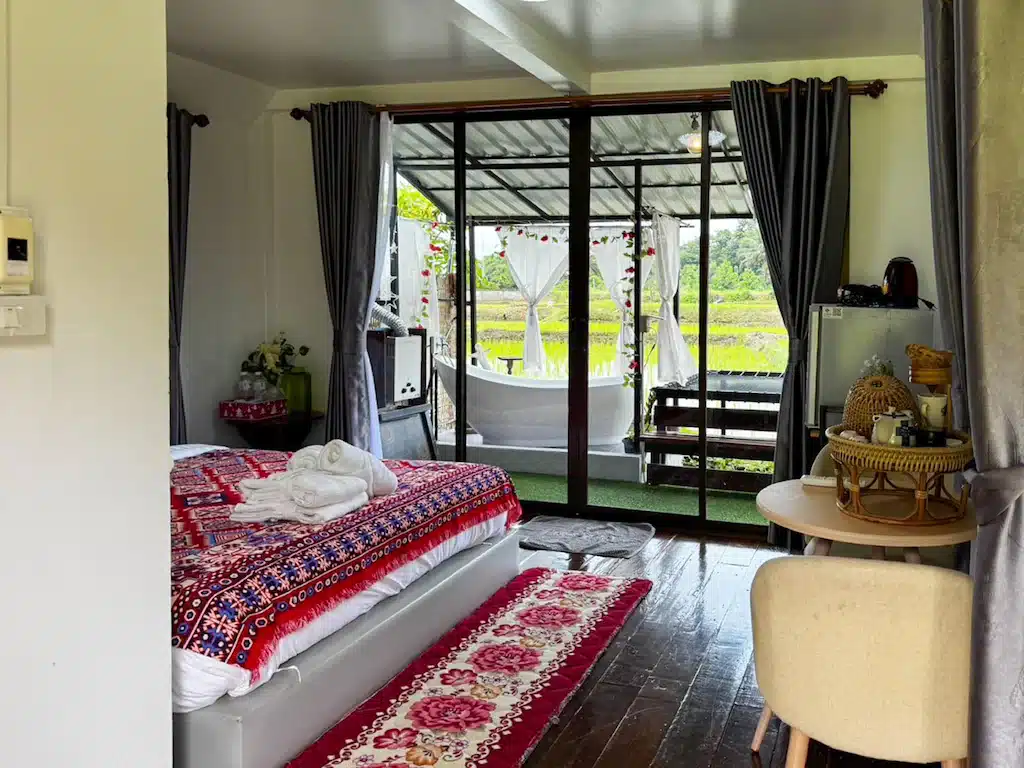 ห้องนอนที่แสนอบอุ่นที่ โรงแรมน่าน พร้อมเตียงที่มีลวดลายสีแดง ผ้าเช็ดตัว โต๊ะรับประทานอาหารขนาดเล็ก และประตูบานเลื่อนกระจกที่เปิดออกสู่ระเบียงพร้อมอ่างอาบน้ำสีขาวและวิวทุ่งหญ้าสีเขียว.