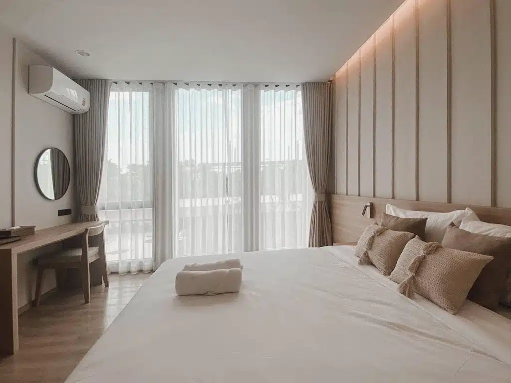 ห้องนอนสไตล์โมเดิร์นมินิมอลที่ ที่พักเชียงใหม่ ประกอบด้วยเตียงขนาดใหญ่ ผ้าปูที่นอนสีกลาง โต๊ะทำงานพร้อมกระจก และหน้าต่างบานใหญ่จากพื้นจรดเพดานพร้อมผ้าม่านบางที่ช่วยให้แสงธรรมชาติส่องเข้ามาได้.