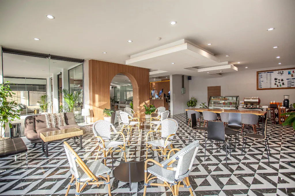 การตกแต่งภายในคาเฟ่สมัยใหม่ด้วยพื้นกระเบื้องลาย, เก้าอี้หวาย, โต๊ะกระจก, โซฟา, พืชเขียวชอุ่ม, และเคาน์เตอร์ที่จัดแสดงขนมอบและเมนู—เหมาะสำหรับแขกจาก โรงแรมเชียงใหม่ ที่ต้องการหาสถานที่อบอุ่นเพื่อผ่อนคลาย.