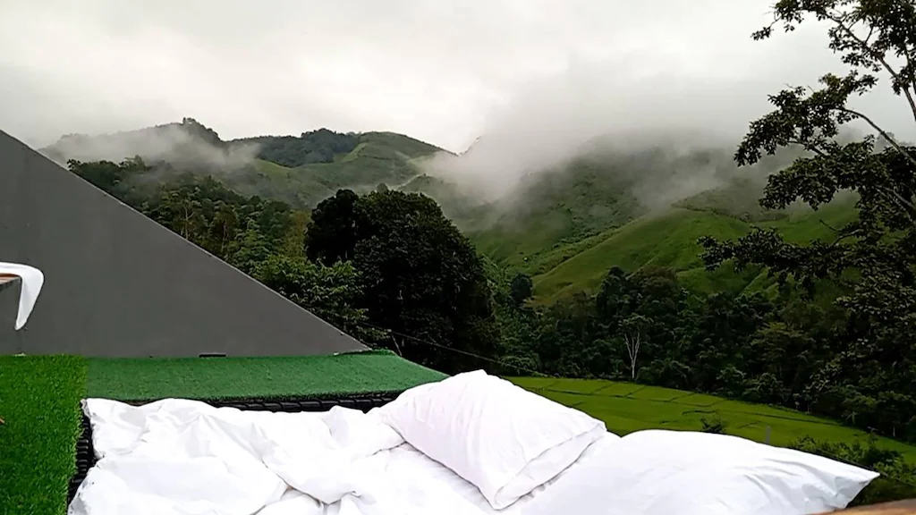 หมอนสีขาวและผ้าห่มบนเตียงกลางแจ้ง มองเห็นเนินเขาสีเขียวและต้นไม้ที่ปกคลุมด้วยหมอก—ภาพที่ชวนให้หลงใหลที่รีสอร์ทในน่านสำหรับผู้ที่มองหาที่พักในน่านท่ามกลางความงดงามของธรรมชาติ.