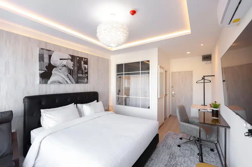 ห้องพักโรงแรมสมัยใหม่ในโรงแรมเชียงใหม่ พร้อมเตียงขนาดใหญ่ โต๊ะทำงาน เก้าอี้ โทรทัศน์ติดผนัง และภาพเหมือนขาวดำเหนือหัวเตียง ห้องตกแต่งด้วยโทนสีกลางและมีฉากกั้นหน้าต่างเพื่อเพิ่มความมีสไตล์.