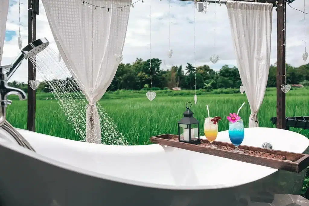 อ่างอาบน้ำกลางแจ้งที่มีน้ำไหลผ่านตั้งอยู่ท่ามกลางทุ่งหญ้าสีเขียวที่รีสอร์ทในน่าน พร้อมเครื่องดื่มสีสันสดใสสองแก้ว โคมไฟ และไฟประดับบนถาดไม้ใต้ผ้าม่านสีขาว.