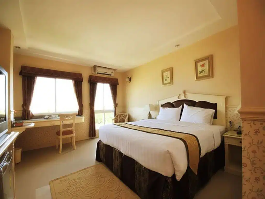 เตียงที่จัดไว้อย่างเรียบร้อยในห้องโรงแรมในเชียงใหม่ที่มีแสงสว่างอบอุ่น หน้าต่างบานใหญ่ โต๊ะและเก้าอี้ โต๊ะข้างเตียง และภาพวาดติดผนัง.