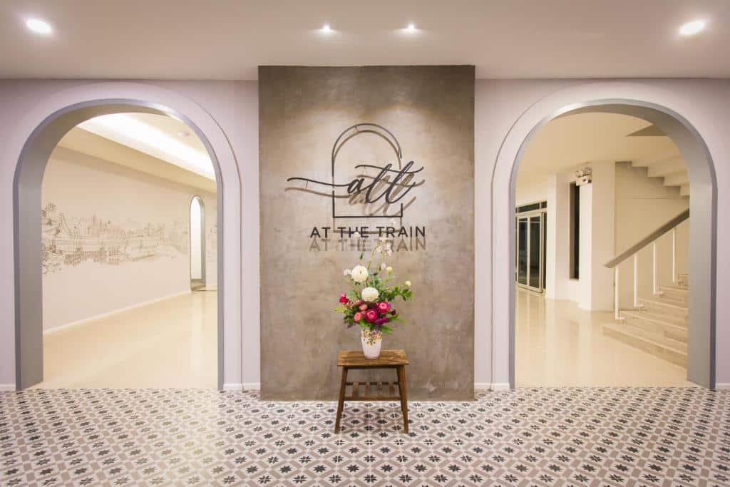 ผนังคอนกรีตกลางที่มีป้าย "At The Train" อยู่ตรงกลาง ขนาบข้างด้วยซุ้มโค้งสองซุ้ม การจัดดอกไม้บนแท่น และกระเบื้องพื้นลายในบรรยากาศภายในที่สว่างไสว—เหมาะสำหรับผู้ที่ต้องการสัมผัสกับบรรยากาศโรงแรมเชียงใหม่ที่ไม่เหมือนใคร.
