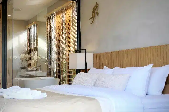 ห้องนอนสมัยใหม่พร้อมเตียงที่จัดไว้อย่างเรียบร้อย หัวเตียงไม้สีอ่อน และม่านบังตาขนาดใหญ่ สามารถมองเห็นอ่างอาบน้ำสีขาวและอ่างล้างหน้าผ่านผนังกระจกได้—เหมาะสำหรับการเข้าพักที่โรงแรมน่านหรือที่พักน่านของคุณ.