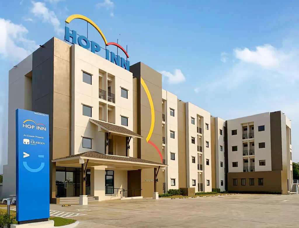 โรงแรมสมัยใหม่สี่ชั้น ภายนอกสีเบจและสีเทา มีป้าย "HOP INN" ขนาดใหญ่บนหลังคา และป้ายสีน้ำเงินที่ทางเข้า—ที่พักในน่านที่เหมาะอย่างยิ่งภายใต้ท้องฟ้าที่มีเมฆบางส่วน.
