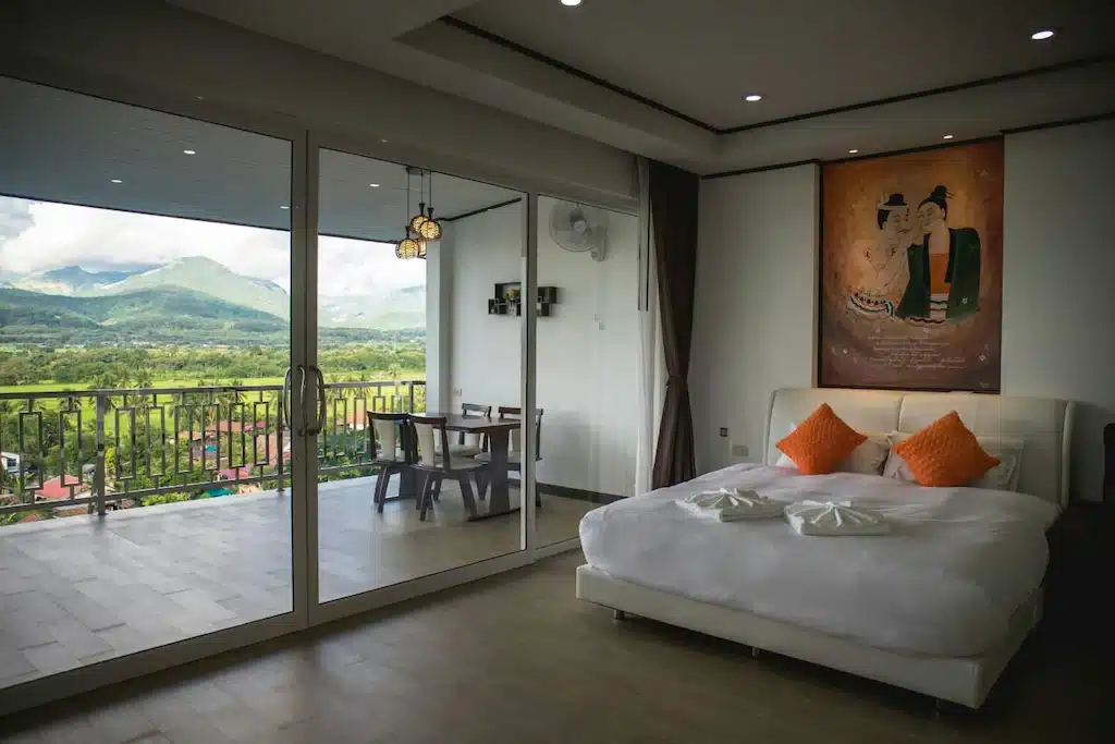 ห้องนอนสมัยใหม่พร้อมเตียงคู่ หมอนสีส้ม ภาพวาดขนาดใหญ่เหนือหัวเตียง และประตูบานเลื่อนกระจกที่เปิดออกสู่ระเบียงซึ่งมีวิวภูเขาและชนบทที่สวยงามที่รีสอร์ท น่าน.