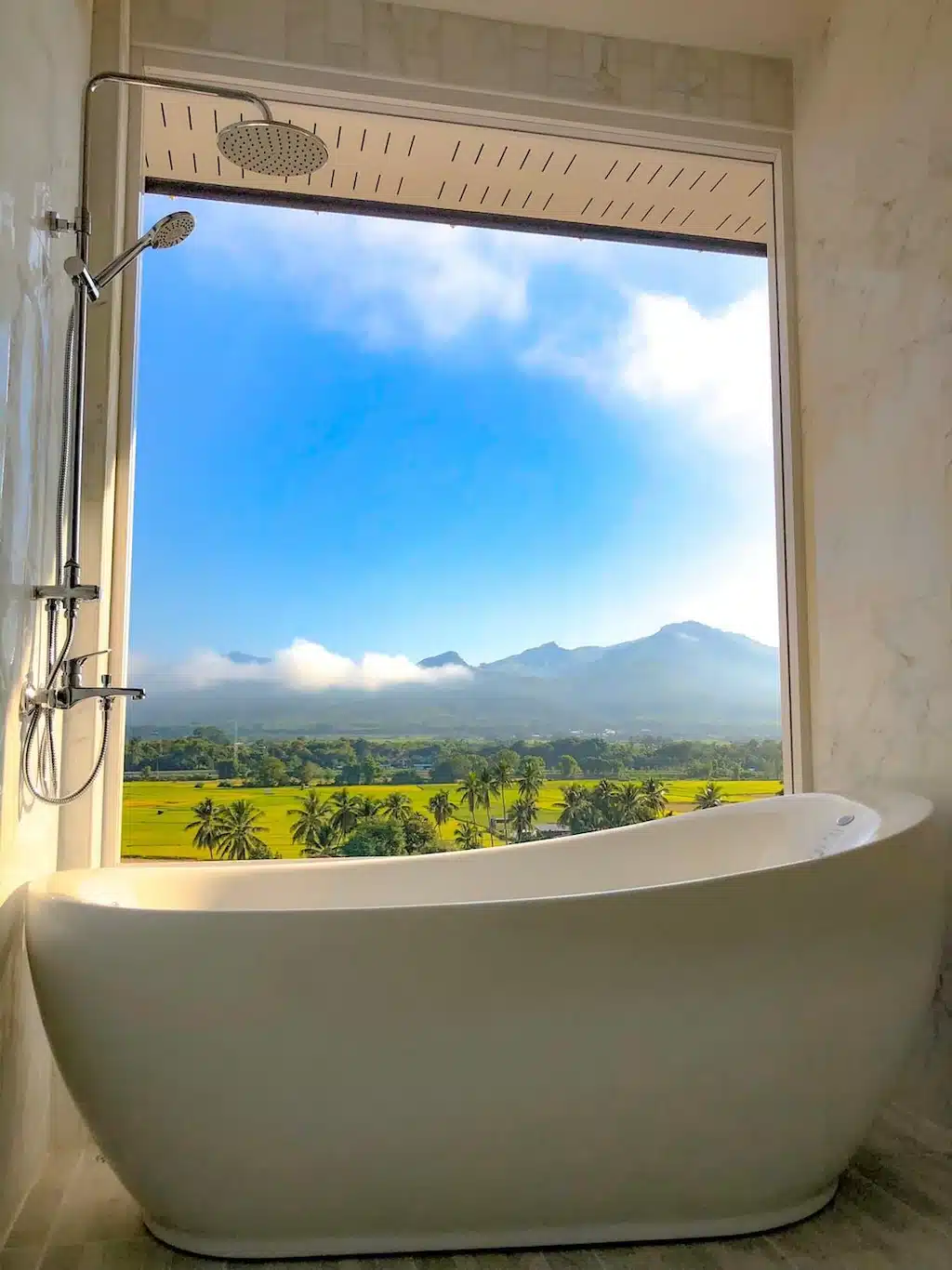 อ่างอาบน้ำสีขาวสมัยใหม่ตั้งอยู่ข้างหน้าต่างบานใหญ่พร้อมฝักบัวเหนือศีรษะ มองเห็นทุ่งนาและภูเขา—เหมาะสำหรับการผ่อนคลายที่โรงแรมน่านพร้อมวิวสวยตระการตาภายใต้ท้องฟ้าสีครามแจ่มใส.