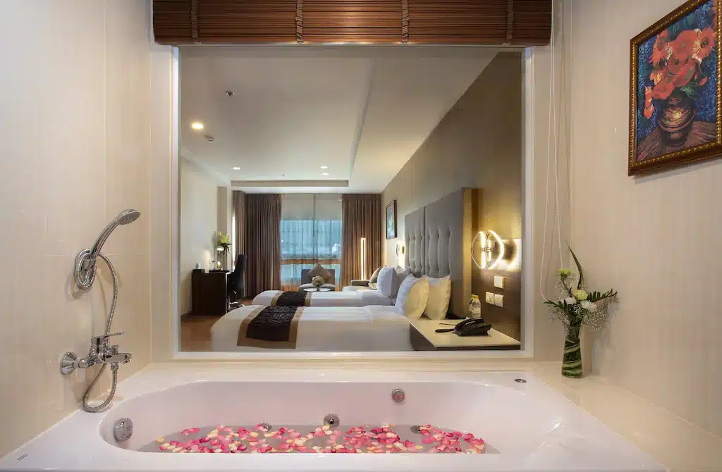 ห้องพักโรงแรมสมัยใหม่ที่ โรงแรมเชียงใหม่, มีอ่างอาบน้ำที่เต็มไปด้วยน้ำและกลีบดอกไม้ในเบื้องหน้า, แยกออกจากห้องนอนด้วยหน้าต่างกระจกขนาดใหญ่.