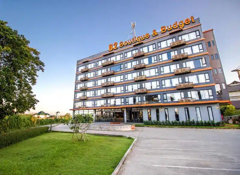 โรงแรมเชียงใหม่ที่ทันสมัยสี่ชั้นพร้อมป้ายที่เขียนว่า "B2 Boutique & Budget" มีระเบียงและลานจอดรถด้านหน้า เป็นโรงแรมเชียงใหม่ที่เหมาะสำหรับนักเดินทางที่มองหาความสะดวกสบายและความสะดวกในการเดินทาง.