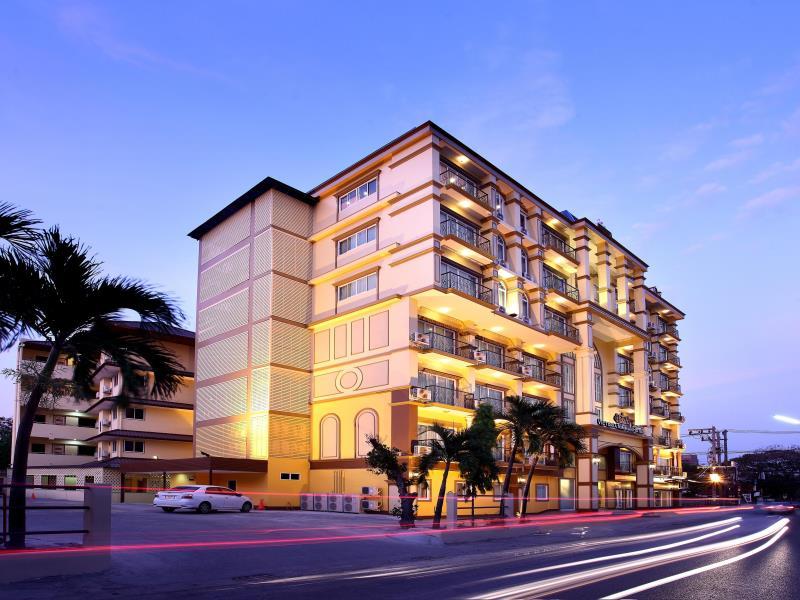 อาคารอพาร์ตเมนต์หลายชั้นสมัยใหม่พร้อมระเบียง ส่องแสงอบอุ่นยามพลบค่ำ ดูคล้ายที่พักเชียงใหม่สุดมีสไตล์ มีต้นปาล์มอยู่ด้านหน้าและเส้นแสงจากรถที่แล่นผ่านไปมาบนถนน.