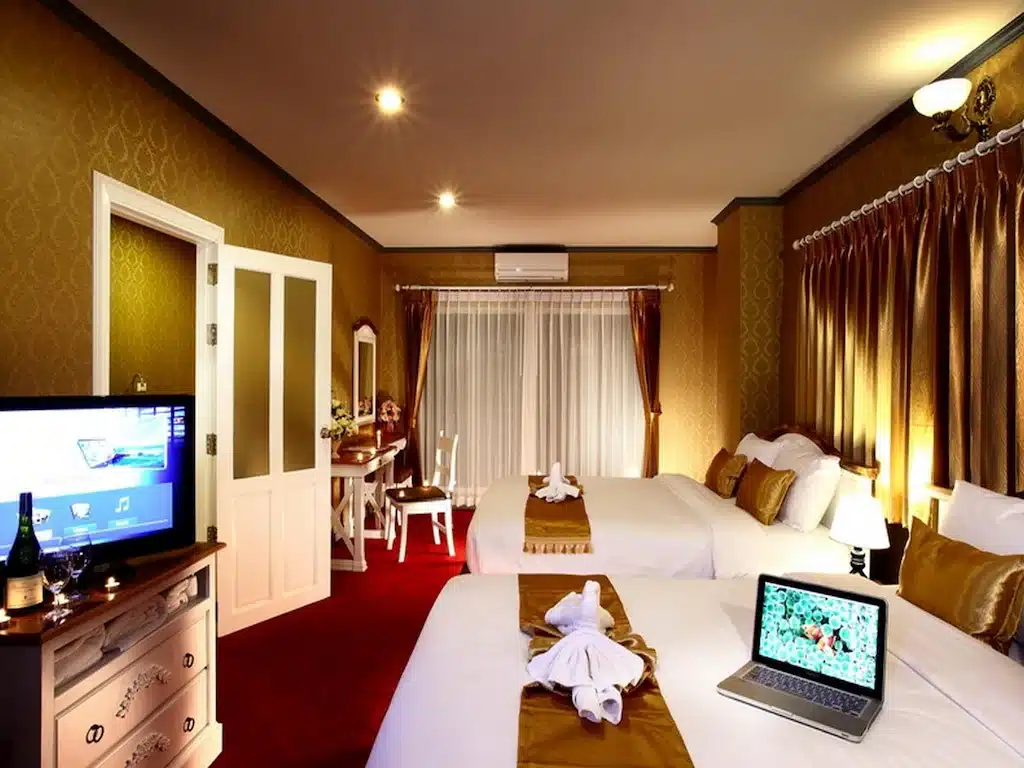 ห้องพักโรงแรมพร้อมเตียงคู่สองเตียง ตกแต่งด้วยสีทอง ทีวี แล็ปท็อปวางอยู่บนเตียงหนึ่ง ผ้าเช็ดตัวจัดเป็นรูปต่างๆ โต๊ะพร้อมกระจก และหน้าต่างพร้อมผ้าม่าน—เหมาะสำหรับการเข้าพักของคุณที่รีสอร์ทเชียงใหม่หรือโรงแรมในเชียงใหม่ที่มีสไตล์.