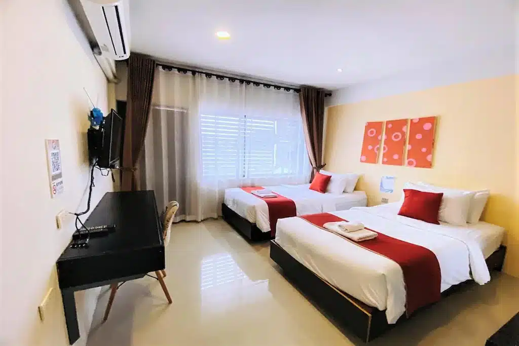 ห้องพักโรงแรมที่ โรงแรมเชียงราย พร้อมเตียงคู่สองเตียง ผ้าปูที่นอนสีแดงและขาว โต๊ะและเก้าอี้ โทรทัศน์ติดผนัง และหน้าต่างบานใหญ่พร้อมผ้าม่าน.