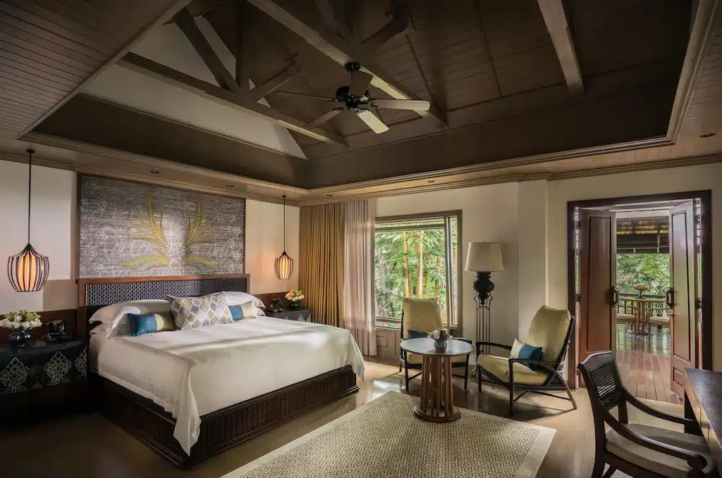 ห้องพักโรงแรมกว้างขวางพร้อมเตียงขนาดใหญ่ พื้นที่นั่งเล่น พัดลมเพดาน และประตูเปิดโล่งสู่ระเบียงที่มองเห็นวิวธรรมชาติเขียวขจี—เหมาะสำหรับการเข้าพักของคุณที่รีสอร์ทเชียงใหม่.
