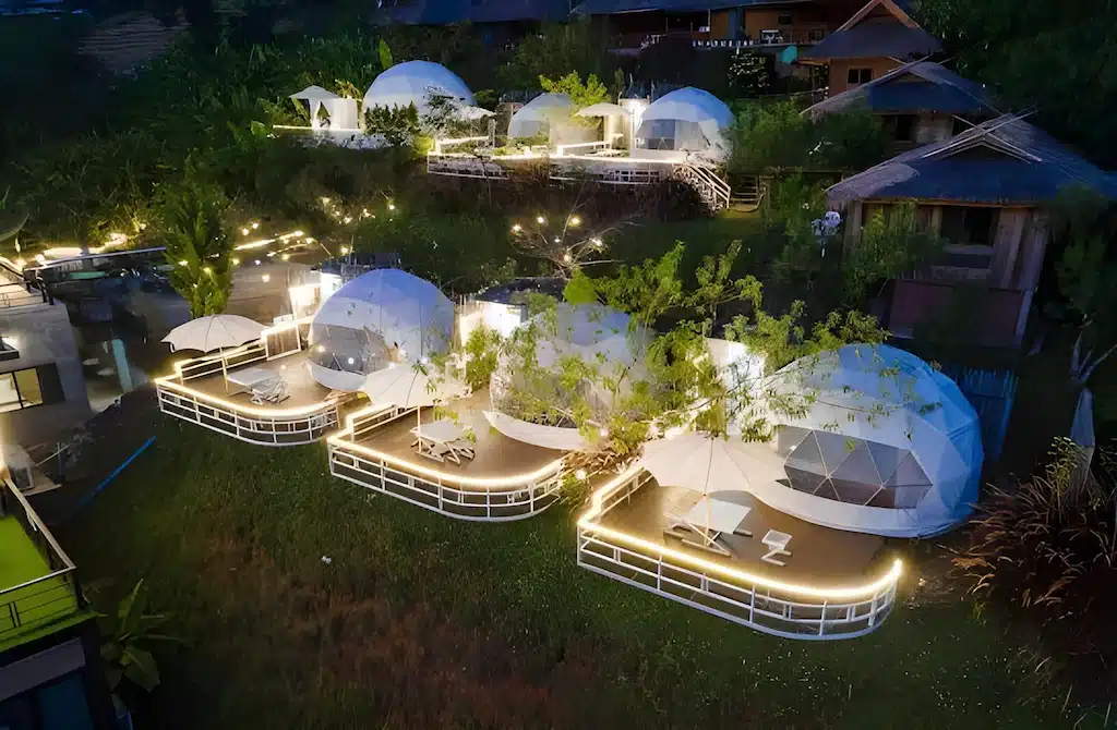 ภาพมุมสูงยามค่ำคืนของเต็นท์โดมจีโอเดสิกหลายหลังที่ส่องสว่างอยู่บนระเบียงของรีสอร์ทเชียงใหม่ ล้อมรอบด้วยพืชพรรณสีเขียวและพื้นที่นั่งเล่นกลางแจ้ง.