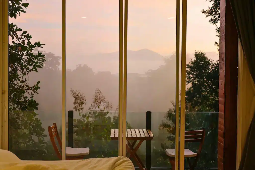 โต๊ะไม้ขนาดเล็กพร้อมเก้าอี้สองตัวตั้งอยู่บนระเบียงที่มองเห็นต้นไม้และภูเขาไกลในยามพระอาทิตย์ขึ้น ผ่านประตูกระจกบานใหญ่—ภาพที่ชวนให้หลงใหลซึ่งเป็นลักษณะเฉพาะของรีสอร์ทที่น่ารักในน่าน.