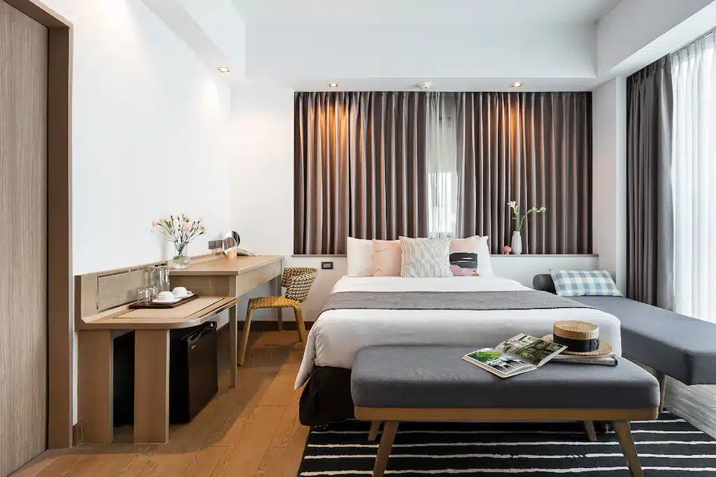 ห้องพักโรงแรมสมัยใหม่ที่ โรงแรมเชียงใหม่ พร้อมเตียงคู่ โต๊ะข้างเตียง โต๊ะทำงานพร้อมเก้าอี้ ม้านั่ง โซฟาเอนนอน และหน้าต่างบานใหญ่ มีนิตยสารและที่วางหมวกบนม้านั่งเพื่อความสะดวกสบายของคุณระหว่างการเข้าพัก.