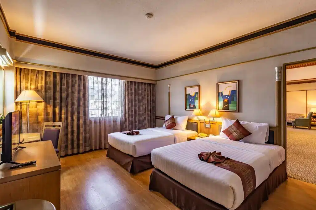 ห้องพักโรงแรมพร้อมเตียงคู่สองเตียง หน้าต่างพร้อมผ้าม่าน โต๊ะทำงาน เก้าอี้ โทรทัศน์ โคมไฟข้างเตียง ภาพศิลปะติดกรอบ และพื้นที่นั่งเล่นเชื่อมต่อ เหมาะสำหรับแขกที่กำลังมองหาที่พักเชียงใหม่เพื่อความสะดวกสบายและความสะดวกในการเข้าพัก.