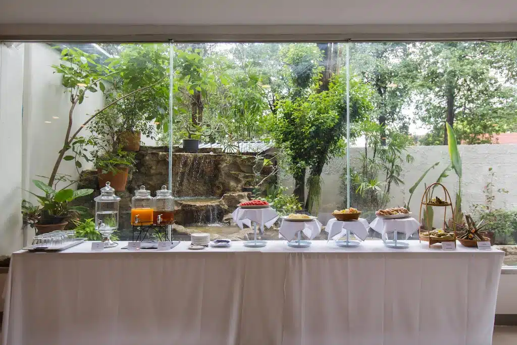 โต๊ะบุฟเฟ่ต์พร้อมเครื่องดื่มและอาหารหลากหลายชนิดถูกจัดเตรียมไว้ภายในอาคารที่รีสอร์ทเชียงใหม่ หน้าต่างบานใหญ่ที่มองเห็นสวนที่มีต้นไม้สีเขียวและน้ำตกขนาดเล็ก.