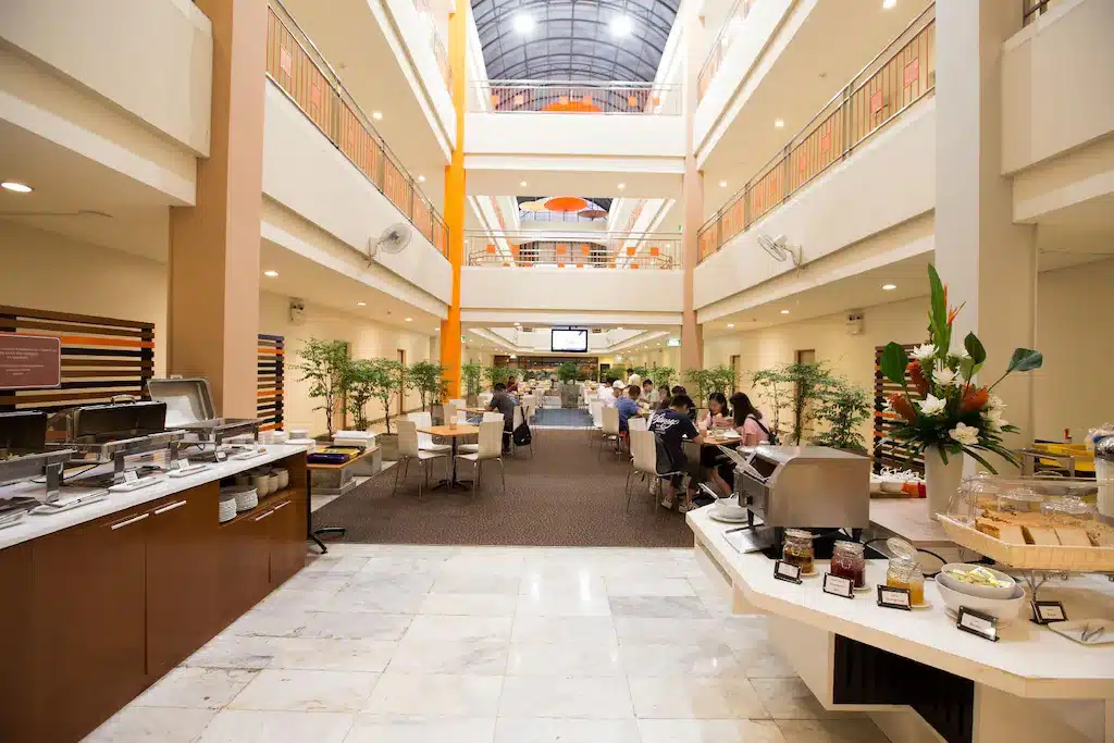 พื้นที่รับประทานอาหารของโรงแรมที่กว้างขวาง ณ โรงแรมเชียงใหม่ พร้อมการจัดเตรียมบุฟเฟ่ต์ไว้ทางซ้ายมือ โต๊ะอาหารพร้อมแขกนั่งรับประทานอยู่ตรงกลาง และแสงสว่างจากธรรมชาติส่องผ่านเพดานกระจกด้านบน.