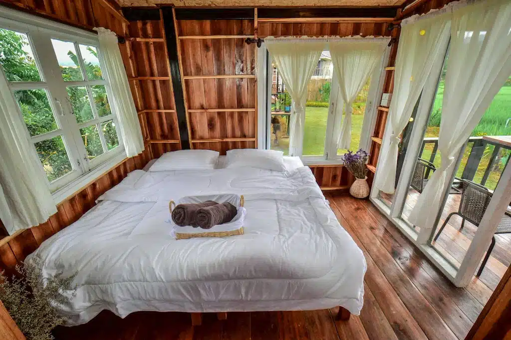 ห้องนอนไม้ที่อบอุ่นที่ ที่พักน่าน พร้อมเตียงคู่ที่ปูผ้าขาวสะอาด, ผ้าเช็ดตัวสองผืนที่ม้วนไว้, และหน้าต่างขนาดใหญ่ที่มองเห็นทิวทัศน์ภายนอกที่เขียวชอุ่ม.