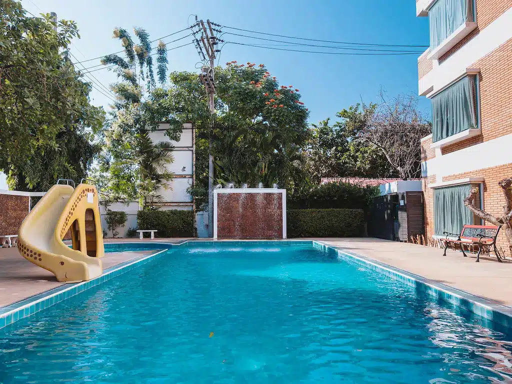 สระว่ายน้ำกลางแจ้งข้างอาคารที่ โรงแรมเชียงใหม่ พร้อมสไลเดอร์สีเหลือง ม้านั่งริมสระ และต้นไม้ล้อมรอบภายใต้ท้องฟ้าสีฟ้าใส.