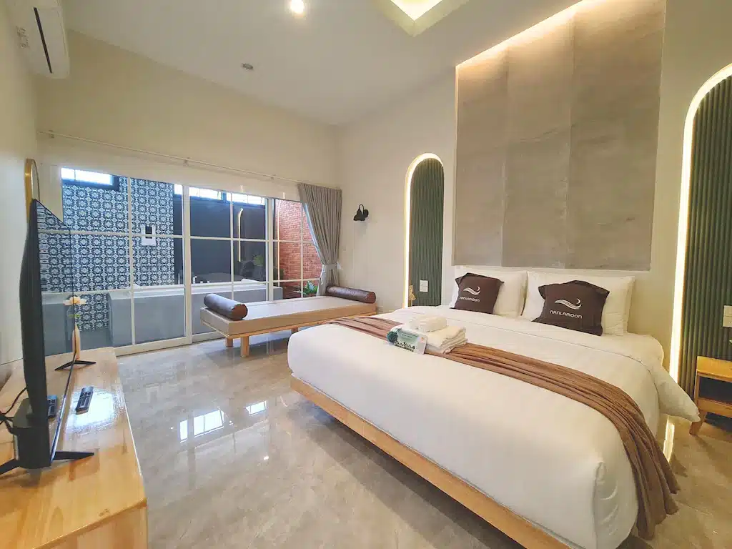 ห้องพักโรงแรมสมัยใหม่พร้อมเตียงขนาดใหญ่, หมอนสองใบ, ม้านั่งข้างหน้าต่าง, โทรทัศน์บนโต๊ะไม้, และพื้นกระเบื้อง; แสงสว่างจากหน้าต่างขนาดใหญ่—เหมาะสำหรับผู้ที่ต้องการโรงแรมในน่านเพื่อการพักผ่อนที่สบาย.