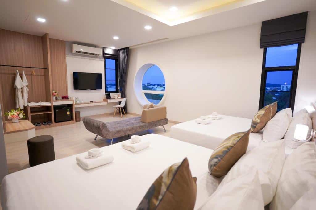 ห้องพักโรงแรมสมัยใหม่พร้อมเตียงคู่สองเตียง, โซฟา, ทีวีติดผนัง, โต๊ะทำงาน, ตู้เสื้อผ้าแบบเปิด, และหน้าต่างกลมที่มองเห็นทัศนียภาพของเมือง—เหมาะสำหรับนักเดินทางที่ต้องการที่พักเชียงรายที่มีสไตล์ในใจกลางเมือง.
