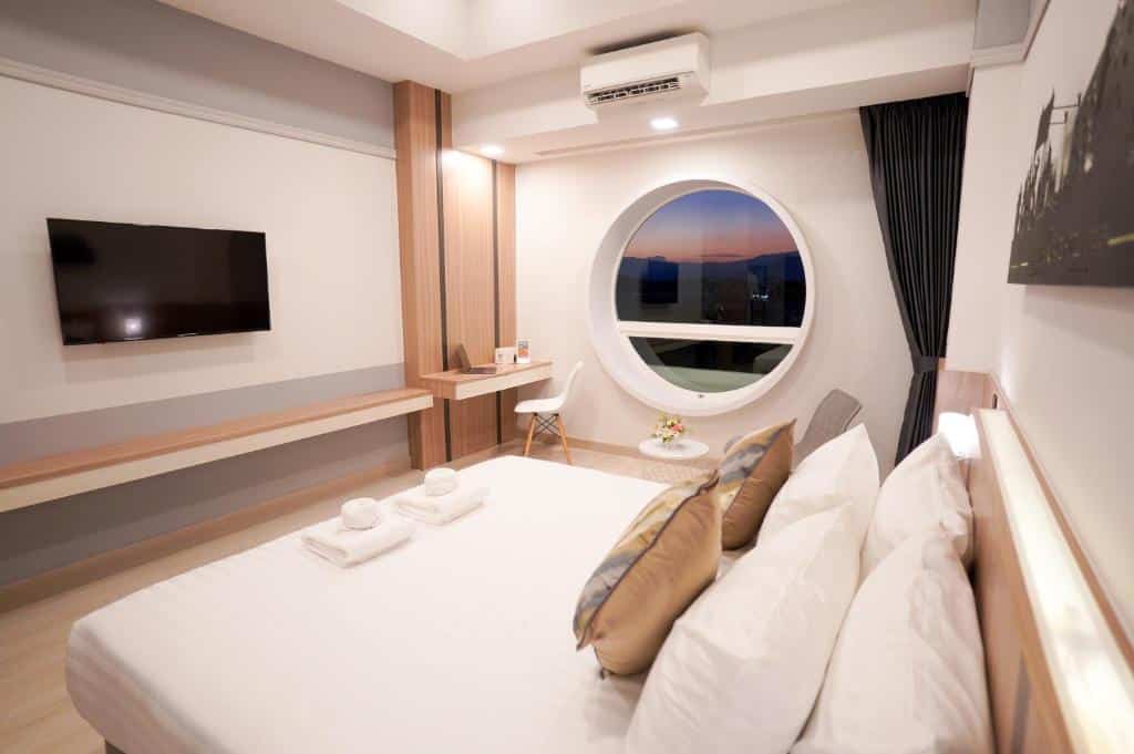 ห้องพักโรงแรมสมัยใหม่พร้อมเตียงขนาดใหญ่ ทีวีติดผนัง โต๊ะทำงาน และหน้าต่างทรงกลมที่มองเห็นวิวพระอาทิตย์ตก ผ้าเช็ดตัวพับไว้บนเตียง การตกแต่งเรียบง่ายและทันสมัย—เหมาะสำหรับการเข้าพักของคุณที่โรงแรมเชียงราย.