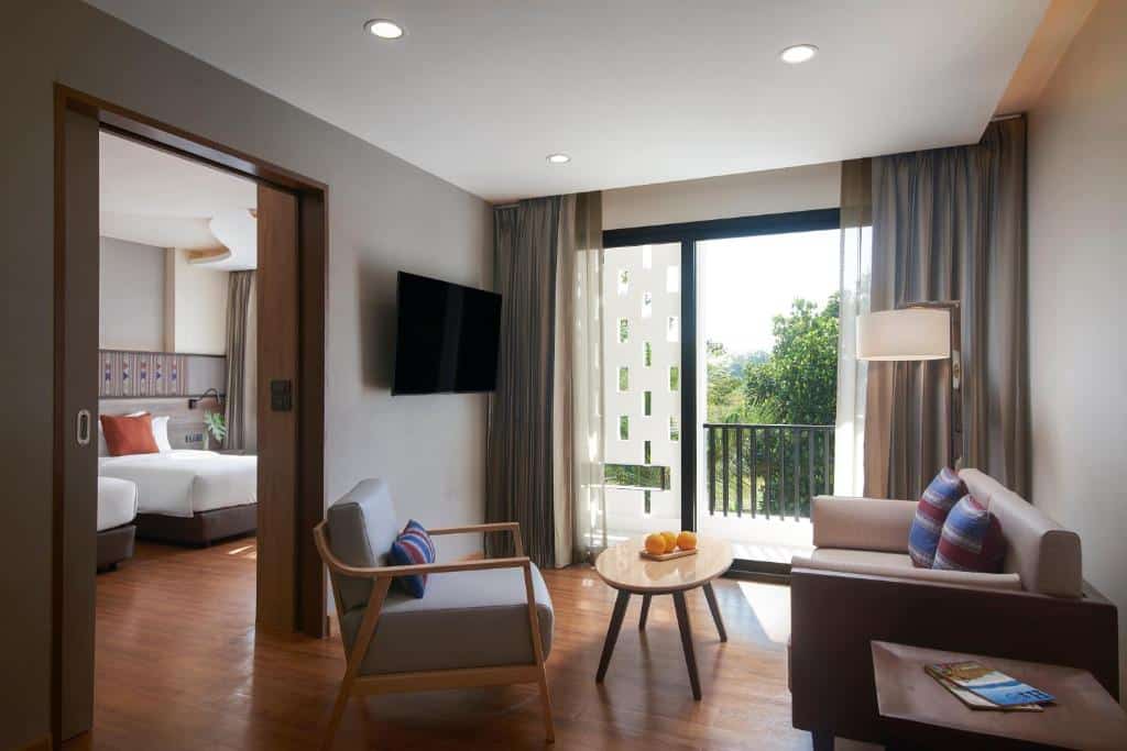 ห้องสวีทโรงแรมสมัยใหม่ใน น่านที่พัก พร้อมห้องนอนแยก ห้องนั่งเล่นขนาดเล็กที่มีโซฟา เก้าอี้นวม และโต๊ะพร้อมผลไม้ รวมถึงระเบียงที่มองเห็นวิวภายนอกที่สวยงาม.