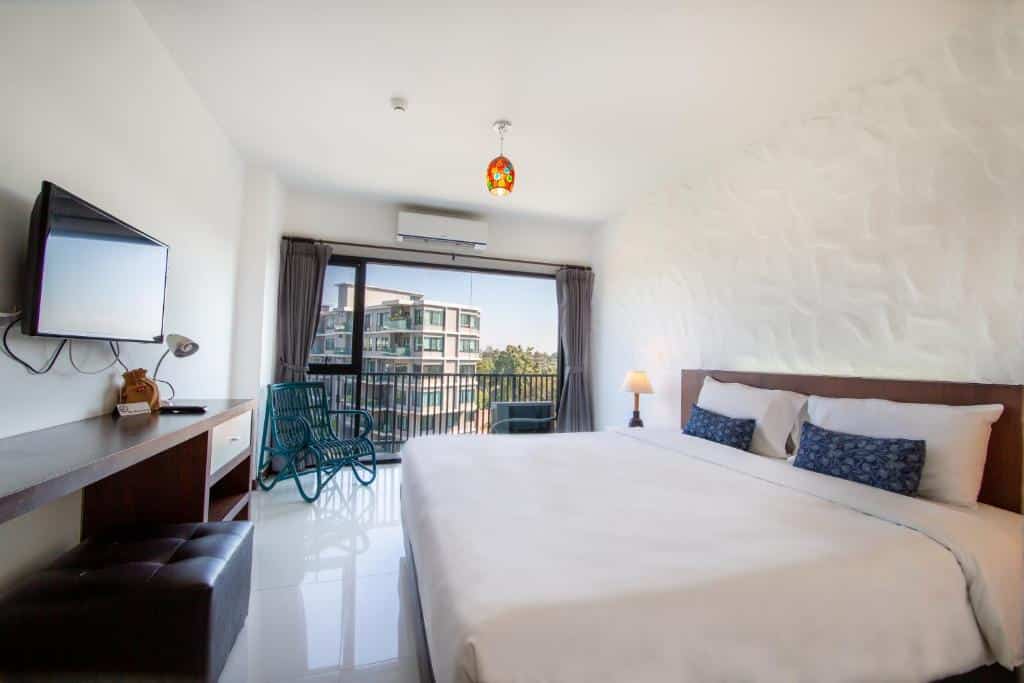 ห้องพักโรงแรมที่สว่างสดใสในโรงแรมเชียงใหม่ พร้อมเตียงขนาดใหญ่ โต๊ะทำงาน โทรทัศน์ และระเบียงที่มองเห็นอาคารและต้นไม้อื่น ๆ แสงธรรมชาติส่องผ่านประตูกระจกบานเลื่อน.