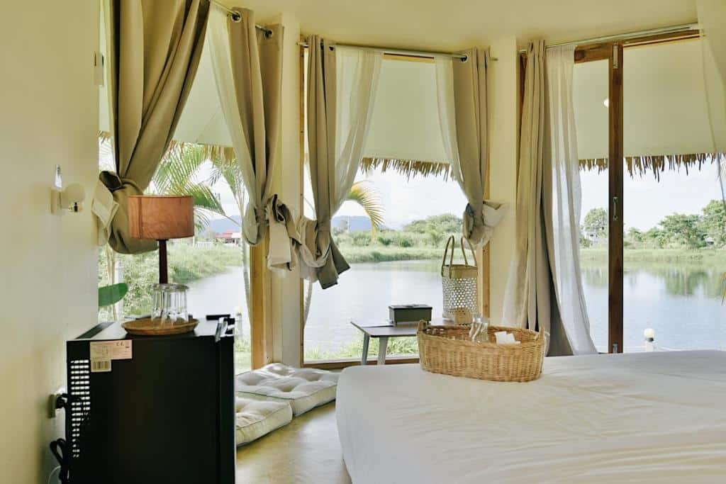 ห้องสว่างไสวที่โรงแรมเชียงราย พร้อมหน้าต่างบานใหญ่ ผ้าม่านบางเบา เตียงนอน ตะกร้าหวาย ตู้เย็นขนาดเล็ก และวิวทะเลสาบและต้นไม้เขียวขจีภายนอก.