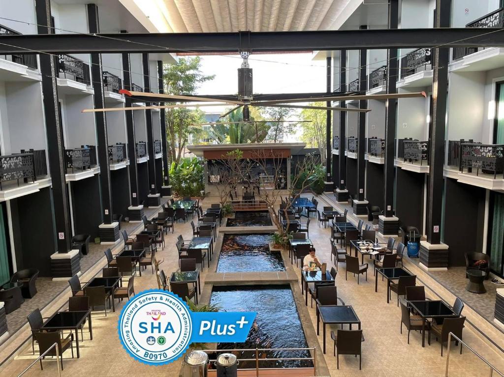 พื้นที่รับประทานอาหารภายในอาคารที่ทันสมัยพร้อมโต๊ะและเก้าอี้สีดำจัดวางรอบบ่อน้ำตกแต่งกลางห้อง ขนาบข้างด้วยระเบียง; ตราสัญลักษณ์รับรองความปลอดภัย SHA Plus+ อยู่ด้านหน้า ณ โรงแรมน่าน เหมาะสำหรับประสบการณ์การเข้าพักในน่านครั้งต่อไปของคุณ.