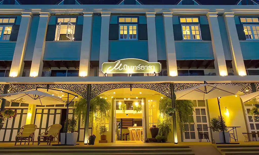 อาคารที่มีแสงสว่างเพียงพอในเวลากลางคืนพร้อมหน้าต่างบานใหญ่ ทางเข้าหลัก ร่มกลางแจ้ง พืช และป้ายไฟที่เขียนเป็นภาษาไทยเหนือประตูทางเข้า—เหมาะสำหรับผู้ที่มองหา โรงแรมน่าน หรือประสบการณ์ที่พักที่ไม่เหมือนใครใน น่าน.