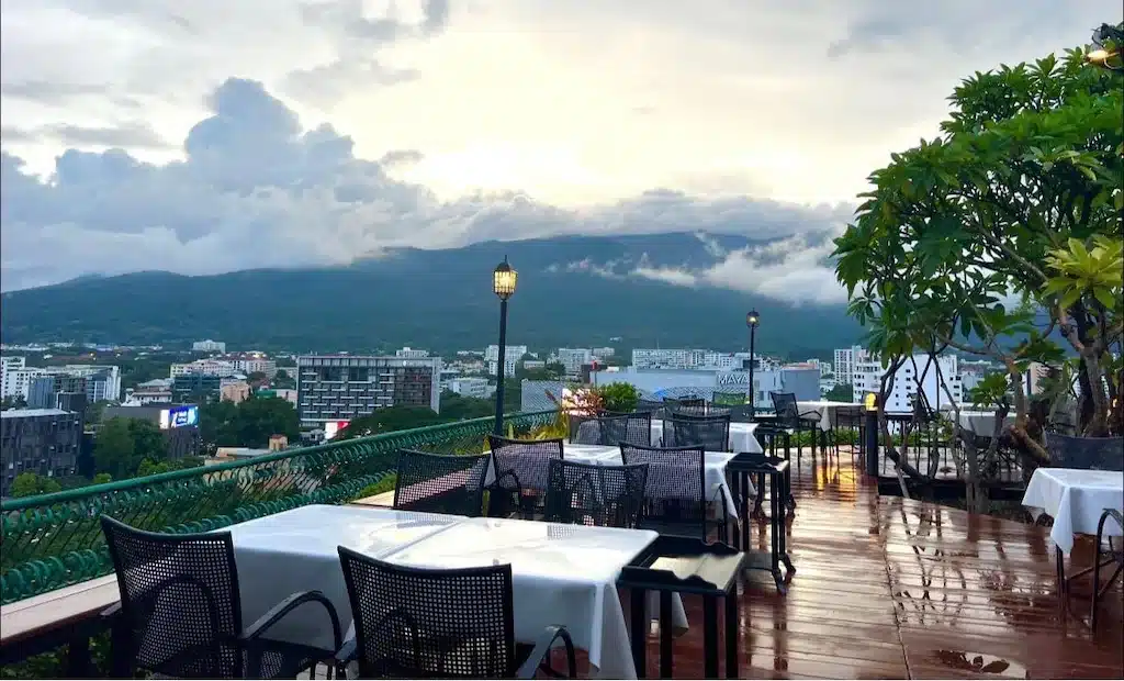 ร้านอาหารบนดาดฟ้าของรีสอร์ทเชียงใหม่ที่มีโต๊ะว่างมองเห็นทิวทัศน์เมือง ภูเขา และท้องฟ้าที่มีเมฆปกคลุม พื้นเปียกบ่งบอกถึงฝนที่เพิ่งตก.